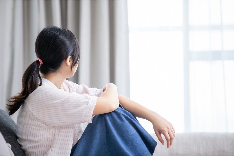 部屋で膝を抱えて窓の外を眺め、婚活の違和感に悩む女性の背中